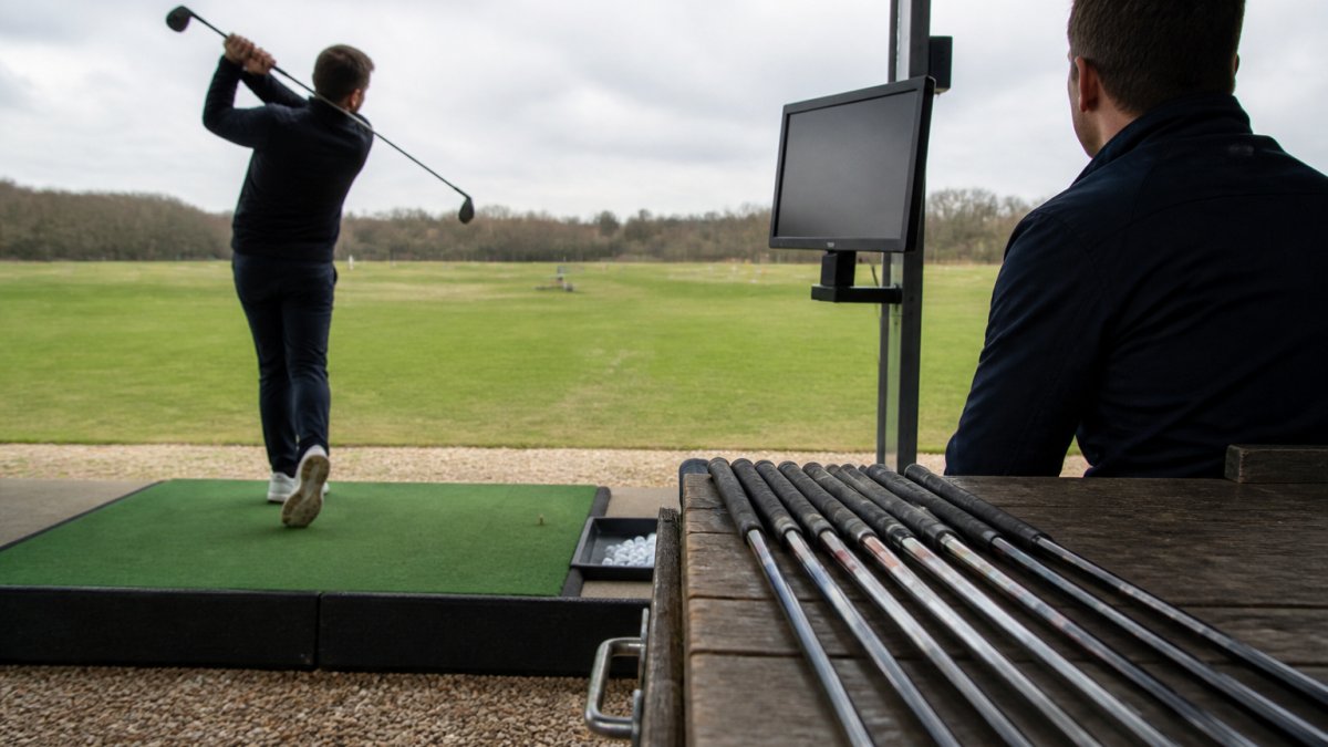 A golfer on a driving range hitting a driver while a fitter watches a launch monitor screen, with several shafts laid out on a bench nearby