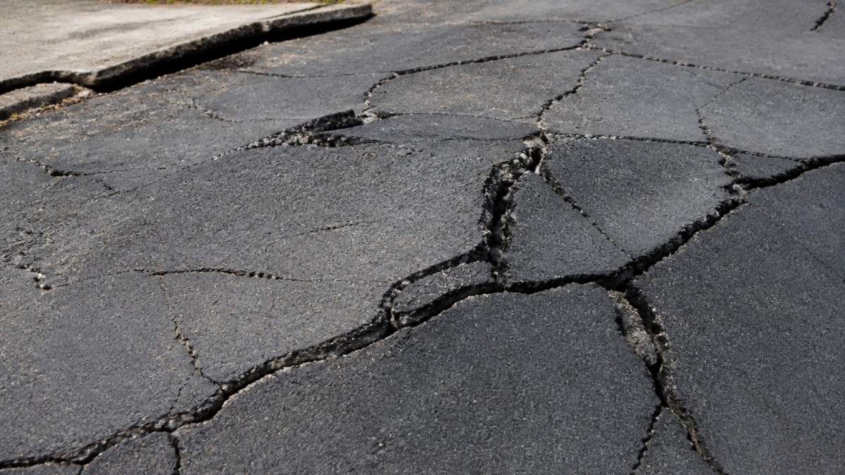 Close-up of severely cracked asphalt driveway surface with heaving and settlement damage visible, showing the impact of expansive clay soil movement underneath