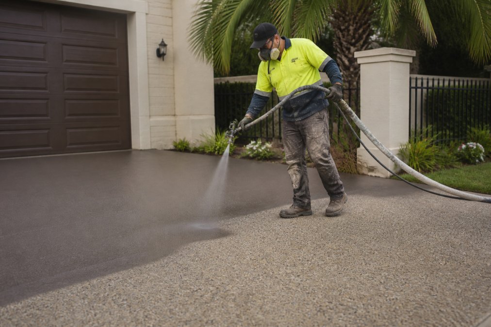 A professional contractor in safety gear using a spray gun to apply a fresh, even coat of decorative concrete resurfacing to a residential driveway, with a finished section visible in the background