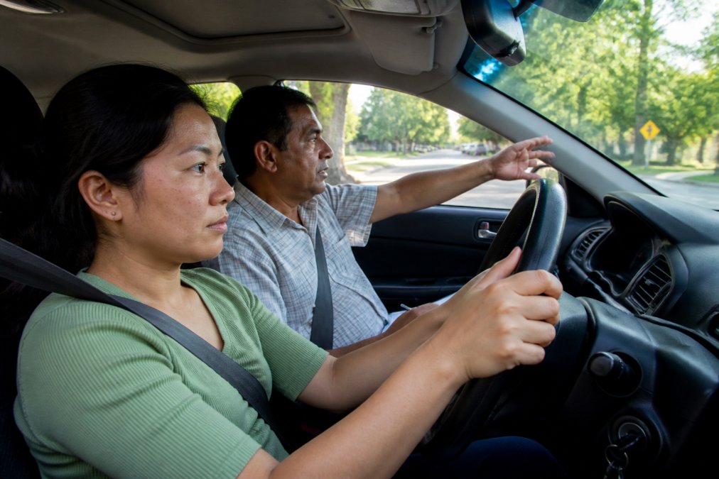 A focused adult learner gripping the steering wheel with calm concentration while a driving instructor gestures toward the road ahead during a lesson on a quiet residential street in soft afternoon light