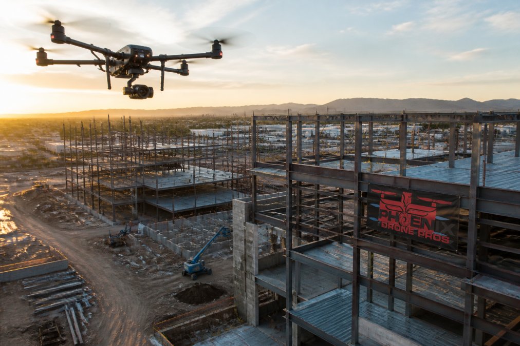 A commercial drone hovering above an active construction site with multiple steel building frames at various stages of completion, shot from a low aerial angle at golden hour, showing the full scale of the development below