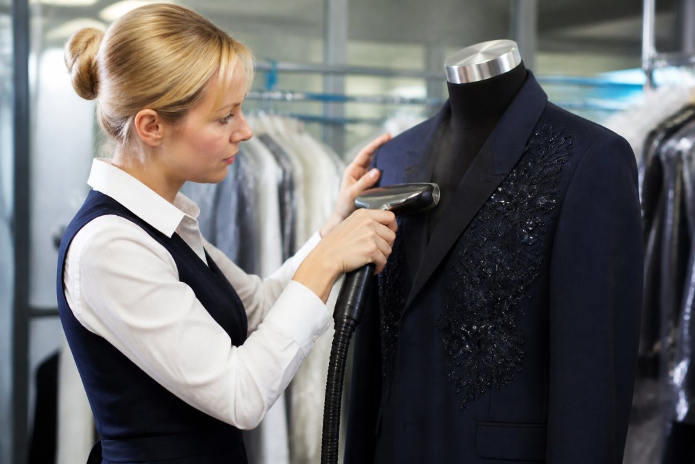 Professional dry cleaner carefully hand-pressing a designer suit jacket with a steamer, showing meticulous attention to embellishments and seams in a well-lit specialty garment care workspace