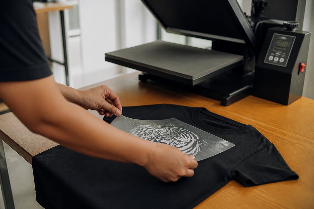 Close-up of hands carefully positioning a DTF transfer onto a black t-shirt, aligning the design precisely before pressing, with a professional heat press visible in the background showing temperature and time controls