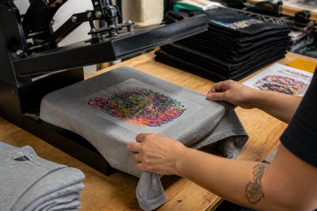 Close-up of hands using a heat press to apply a colourful DTF transfer onto a cotton t-shirt on a workbench, with a stack of finished garments and transfer sheets visible in the background