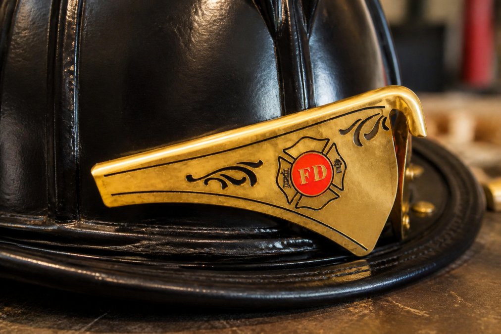 Close-up of a polished brass front holder mounted on a leather fire helmet on a workbench, showing engraved detail and rivet hardware against the helmet shell under warm workshop lighting