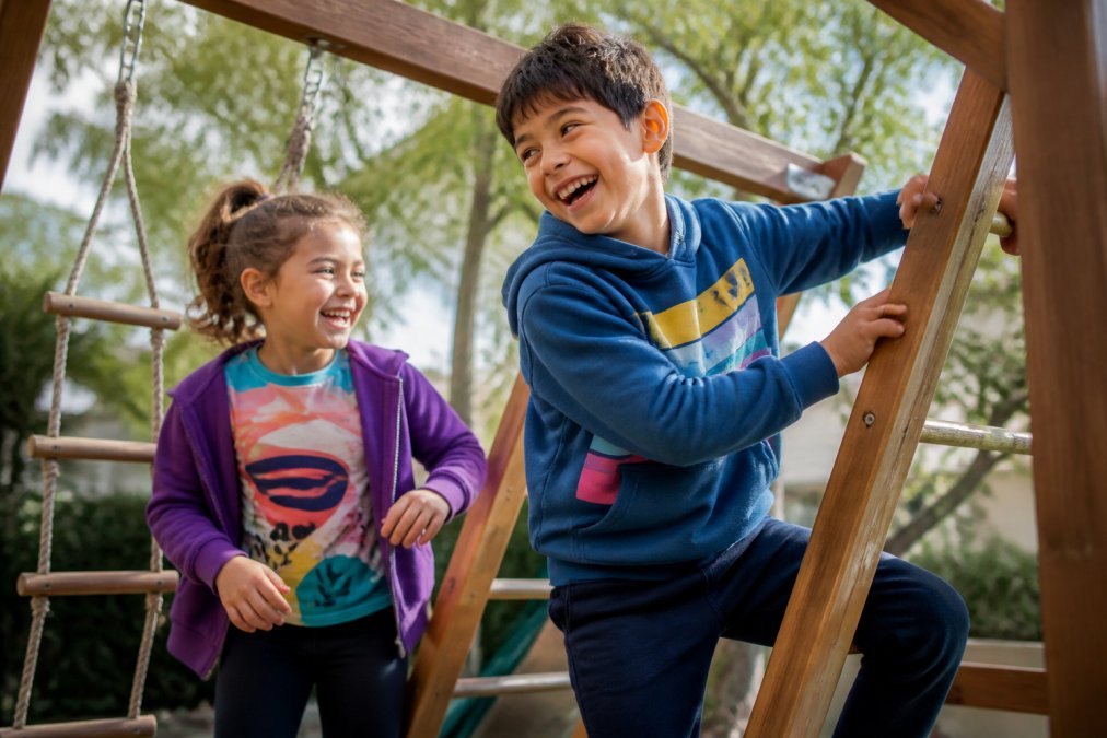 Two active kids, around ages 5 and 9, wearing colorful graphic tees and hoodies while playing on a backyard climbing frame, bright daylight, candid laughter, casual outdoor setting