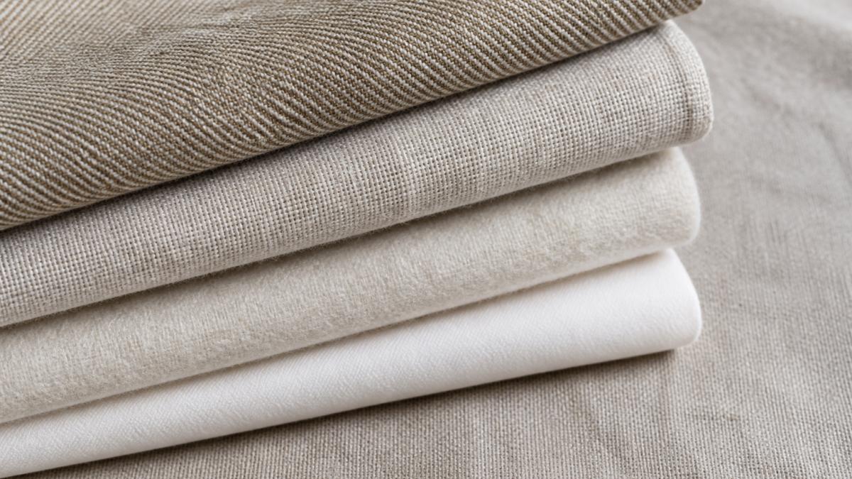 Close-up of folded fabric swatches in different natural fibers showing distinct weave patterns and textures, hemp with tight diagonal weave, linen with crisp structure, merino wool with soft pile, and organic cotton with smooth finish, arranged on a neutral linen background with natural window lighting