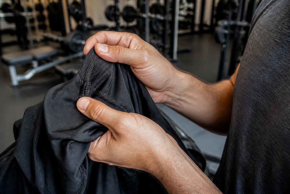 Close-up of a person's hands inspecting the stitching and fabric of a black athletic shirt, with a gym background slightly blurred