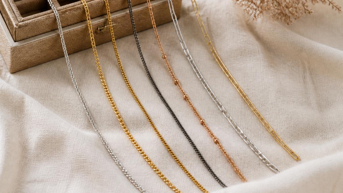 Overhead flat-lay of six necklaces in different metals (silver, gold, dark gray, warm bronze) arranged in a fan on a cream linen cloth beside a small jewelry box