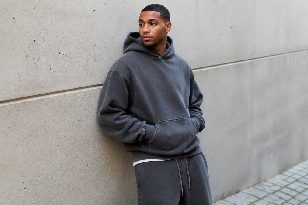 A person wearing an oversized heavyweight cotton hoodie with a relaxed fit, styled casually against a concrete wall in natural daylight, the fabric's thick texture and relaxed silhouette clearly visible