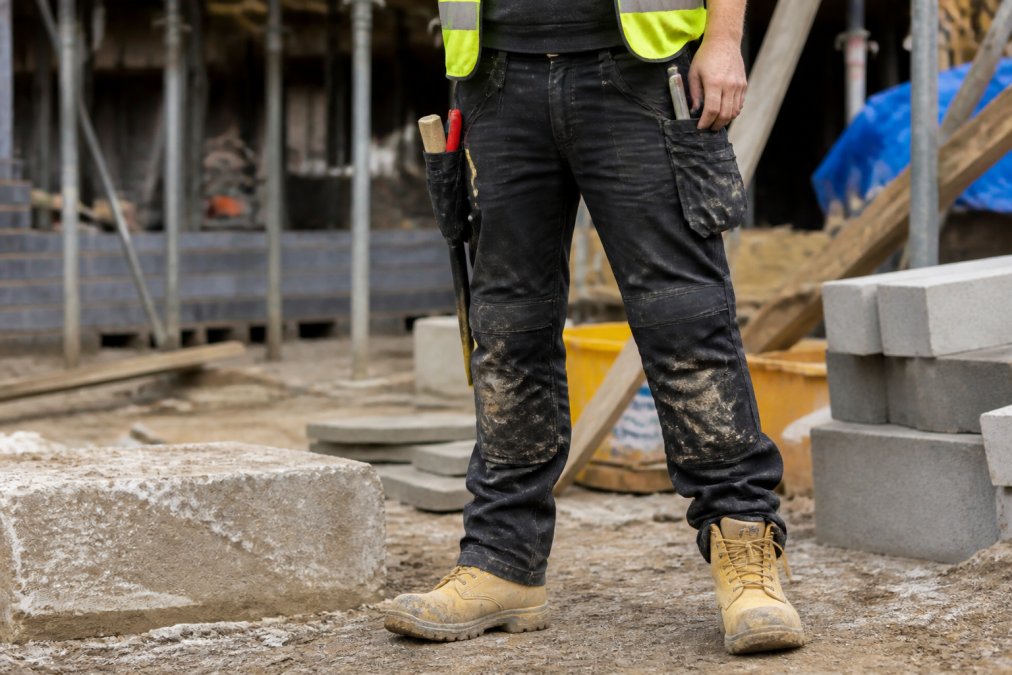 A construction worker wearing black durable work trousers with reinforced knees, standing on a building site with tools in the pockets