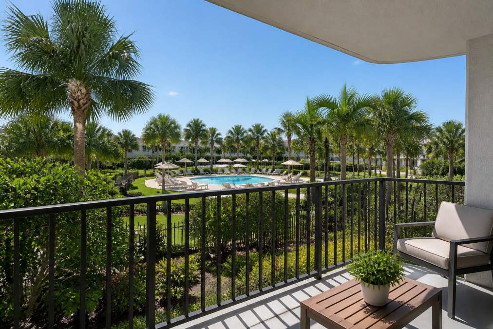Modern apartment balcony overlooking lush landscaped gardens with a resort-style pool in the background, palm trees, and clear blue sky
