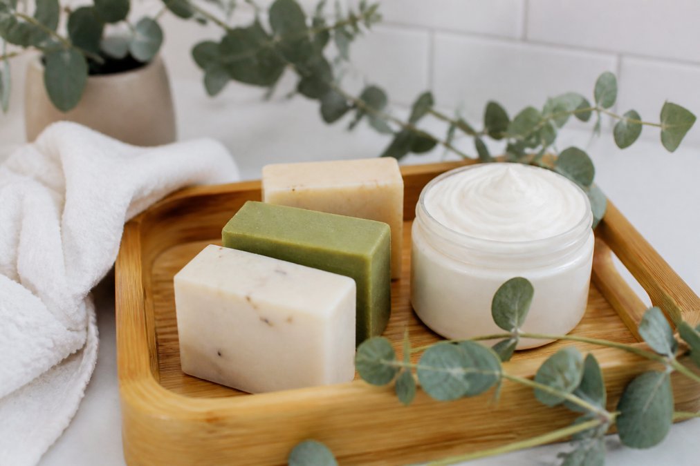 A flat lay of natural, handcrafted soap bars and a whipped body wash jar on a bamboo tray with eucalyptus sprigs and a white towel, bright bathroom lighting