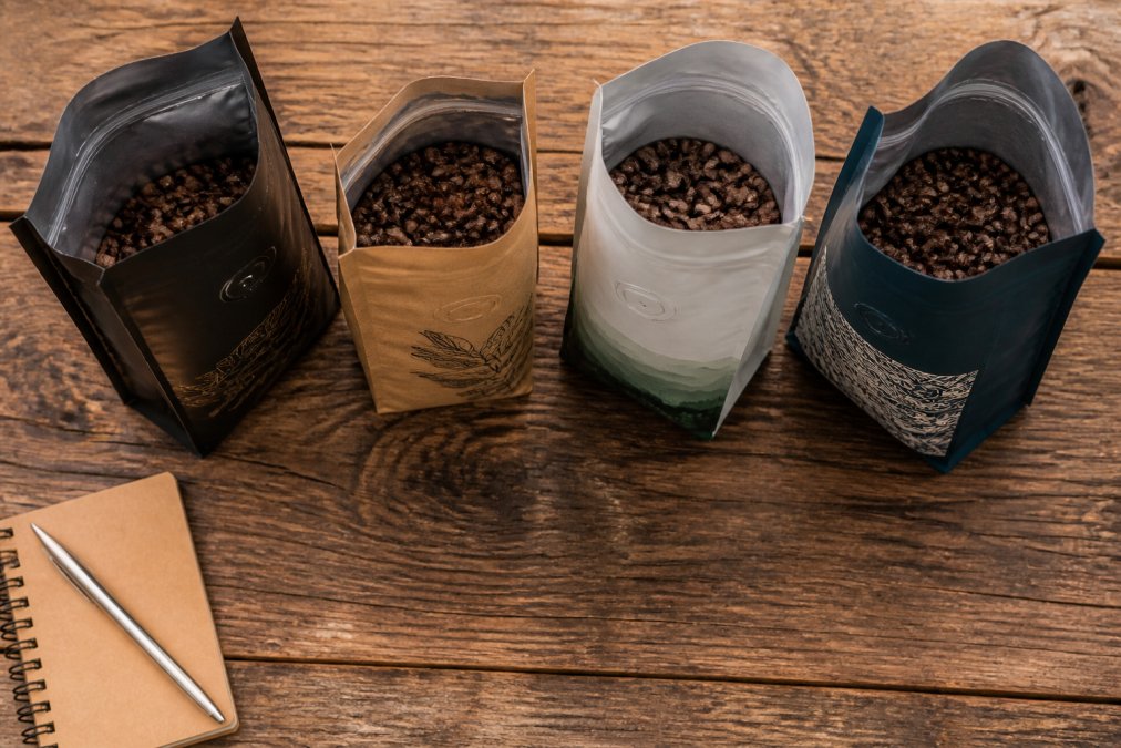 An overhead shot of four different open coffee bags on a wooden table, each with distinct branding and whole beans visible, with a small notebook and pen at the side