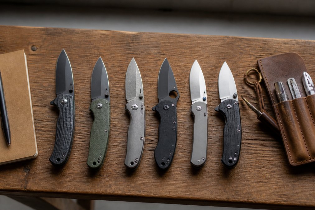 Overhead flat lay of six different folding pocket knives arranged in a row on a wooden workbench, with a pocket notebook and a leather multitool pouch nearby