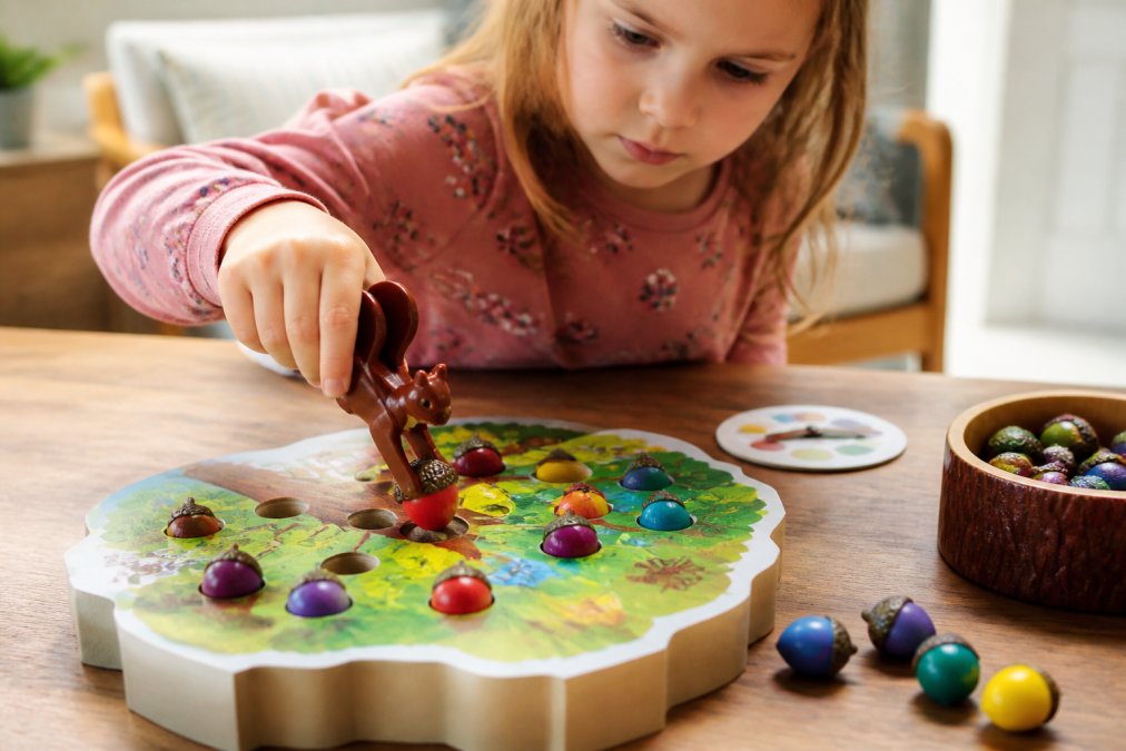 A young child's hands using the squirrel-shaped squeezer to pick up a red acorn from the game board, focused expression, warm natural light on a wooden table at home