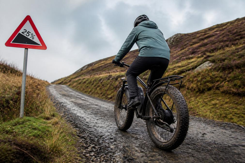 A cyclist in a waterproof jacket riding a fat tyre electric bike up a steep countryside hill past a gradient warning sign, overcast morning light, mud-spattered tyres