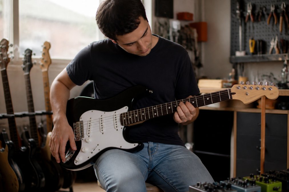 Beginner guitarist examining the neck and fretboard of an electric guitar under natural light, hands positioned to check playability and body comfort against their torso