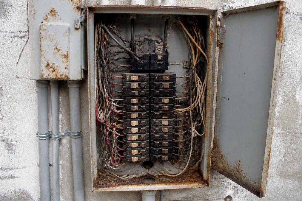 Close-up of an older residential electrical panel with visible rust, crowded wiring, and a mix of mismatched circuit breakers inside an open panel door