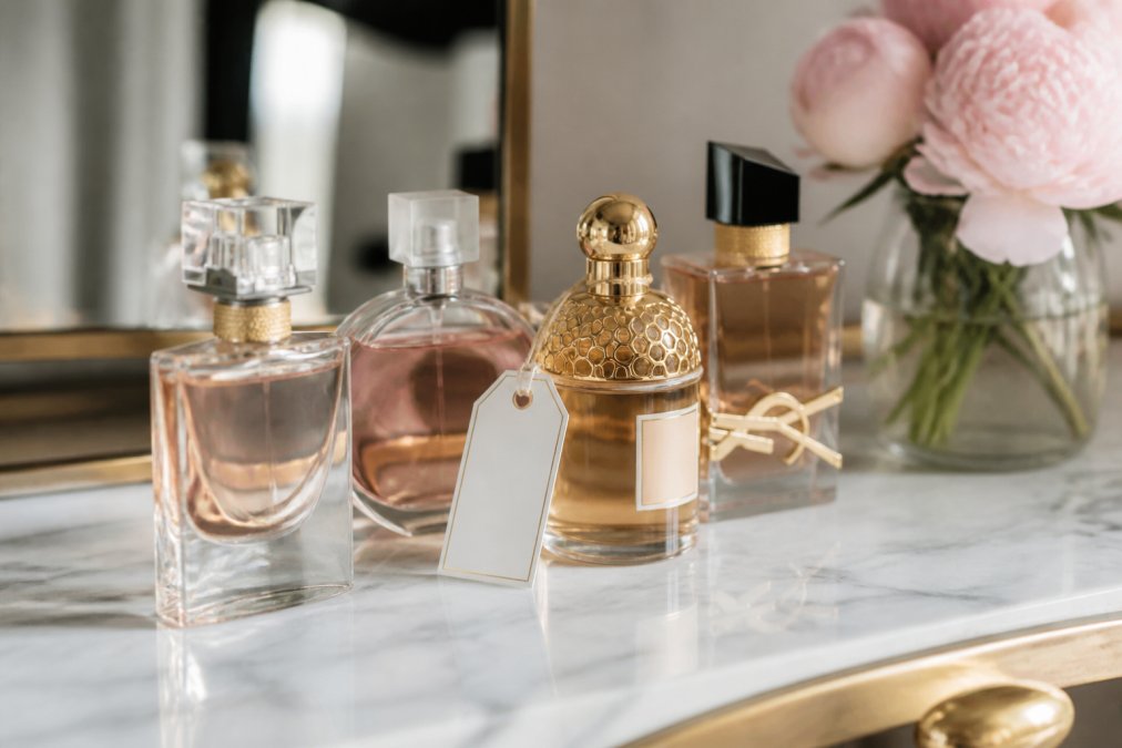 Close-up of several elegant perfume bottles arranged on a marble vanity with soft natural light, a small vase of fresh peonies nearby, and a handwritten gift tag resting against one bottle