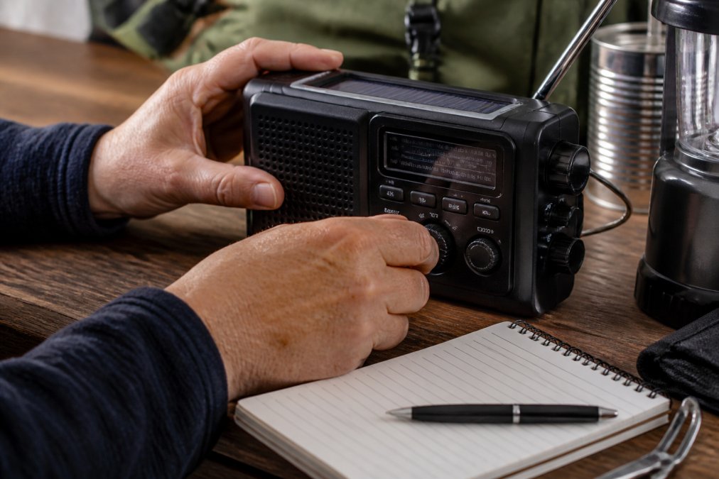 Close-up of hands adjusting an emergency radio's dials and buttons, with a backlit display visible and a notebook for tracking alerts nearby