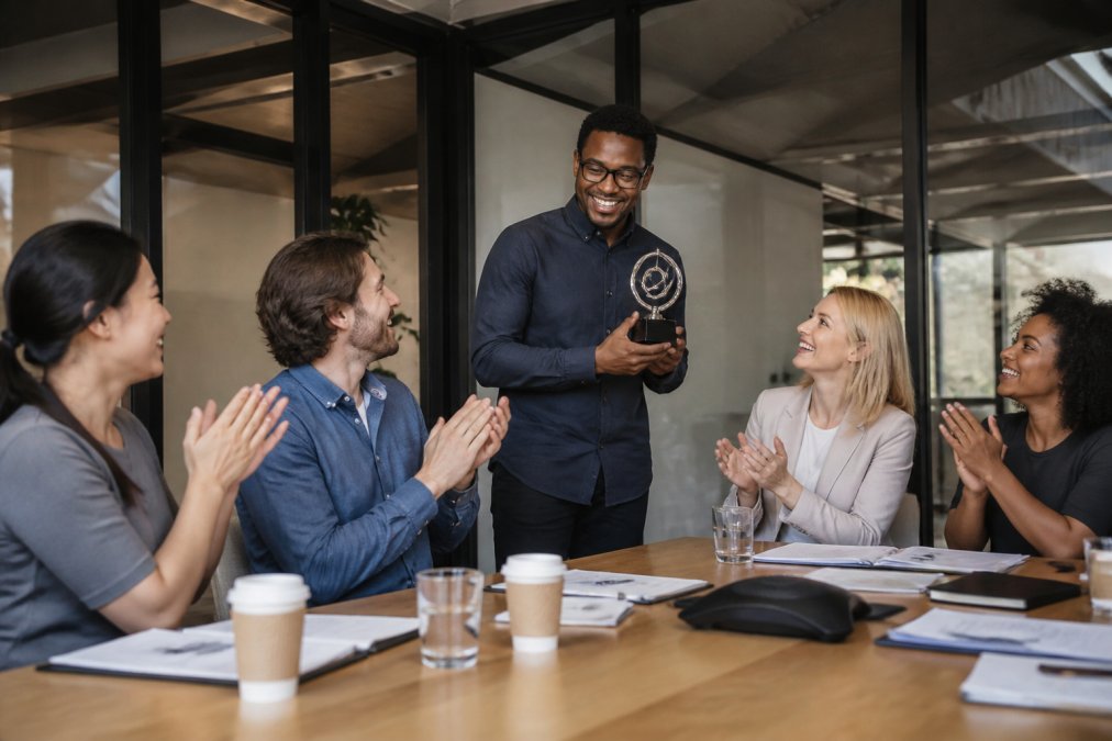A diverse group of employees in a modern office celebrating a colleague's achievement with smiles and applause around a conference table during a team meeting