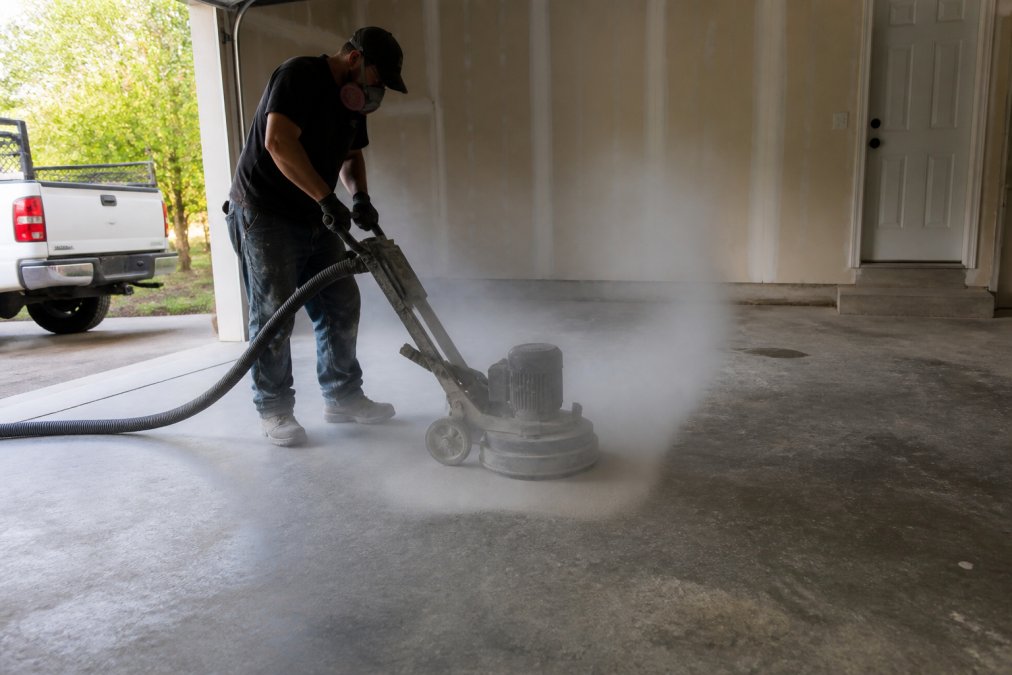 Professional technician operating concrete grinding equipment on residential garage floor, creating dust cloud and exposing prepared substrate ready for epoxy primer application