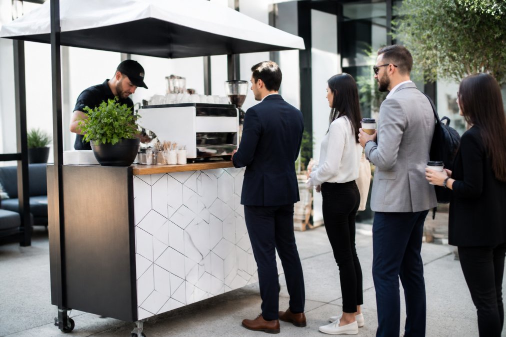 Professional barista preparing specialty espresso drinks at a branded mobile coffee cart with custom logo wrapping and corporate event attendees in business casual attire waiting in line