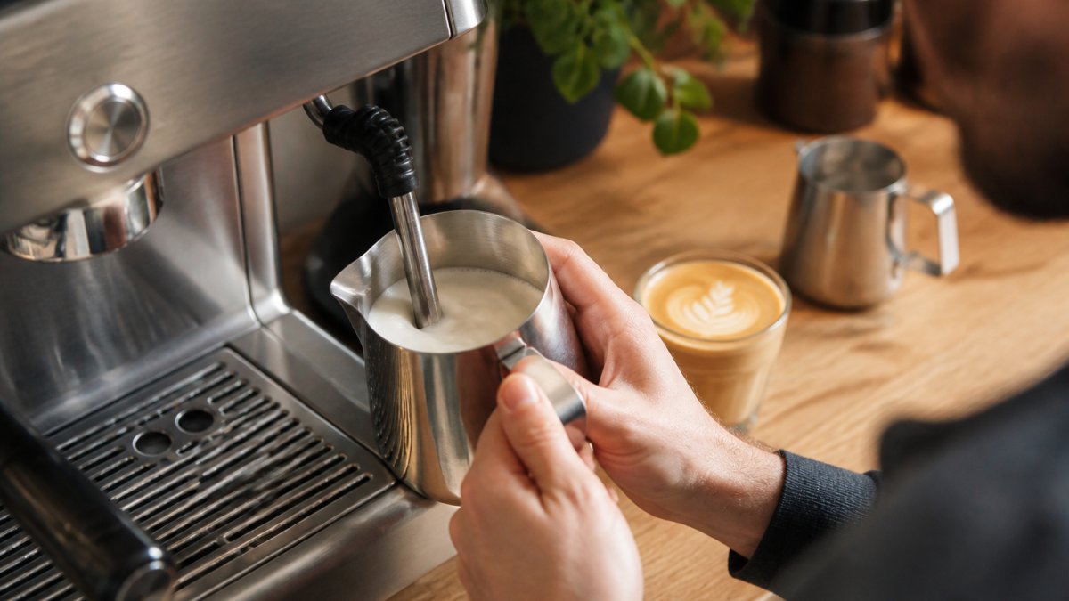 Close-up of a stainless steel milk jug held under a steam wand, glossy microfoam swirling, espresso machine and latte glass on a wooden kitchen counter