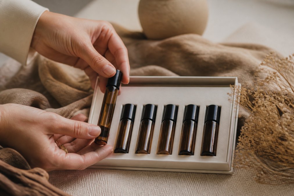 Close-up of fragrance discovery set with six small glass vials arranged on a neutral surface, hands gently holding one vial, natural diffused lighting highlighting the amber glass and product labels