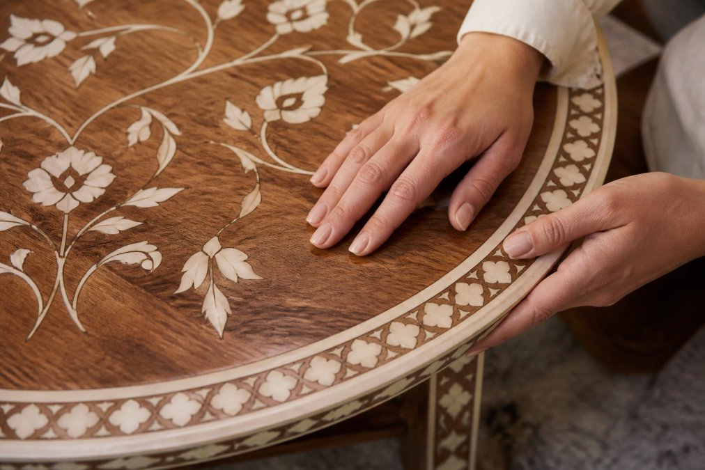 Close-up of hands examining handcrafted wood grain and bone inlay details on a coffee table in natural light, showing the artisan's attention to material character