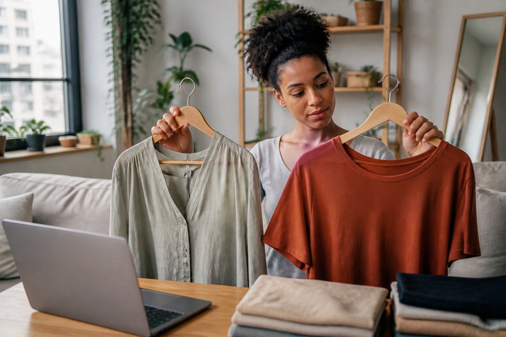 A stylish woman in a bright apartment holding two clothing items on hangers, comparing them thoughtfully, with a laptop open to an online store in the background