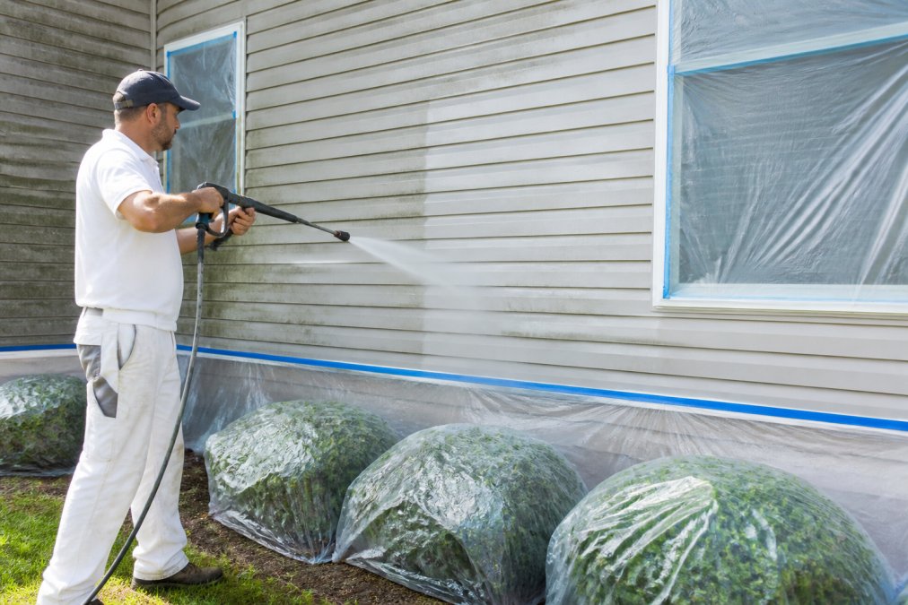Professional painter power washing a residential home's exterior siding with a pressure washer, protective coverings on landscaping and windows, bright daylight conditions