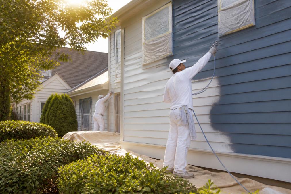 Professional exterior house painters applying fresh paint to residential home siding with professional spray equipment and drop cloths protecting landscaping, morning sunlight