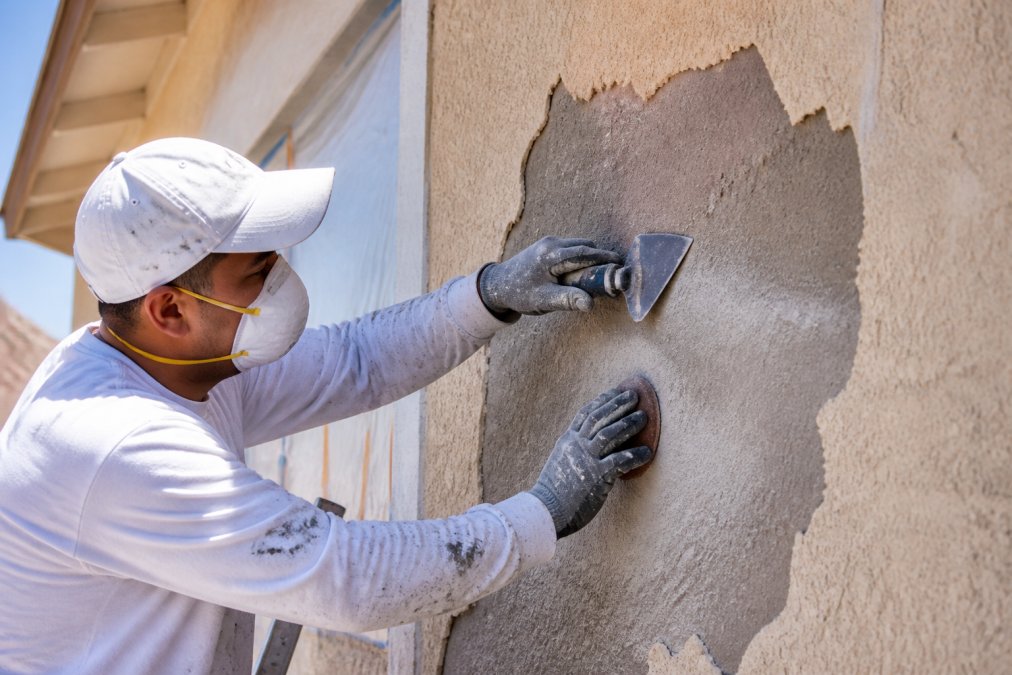 Professional painter scraping and sanding exterior stucco wall surface with handheld tools, showing dust and preparation work in bright daylight, revealing exposed substrate beneath failing paint