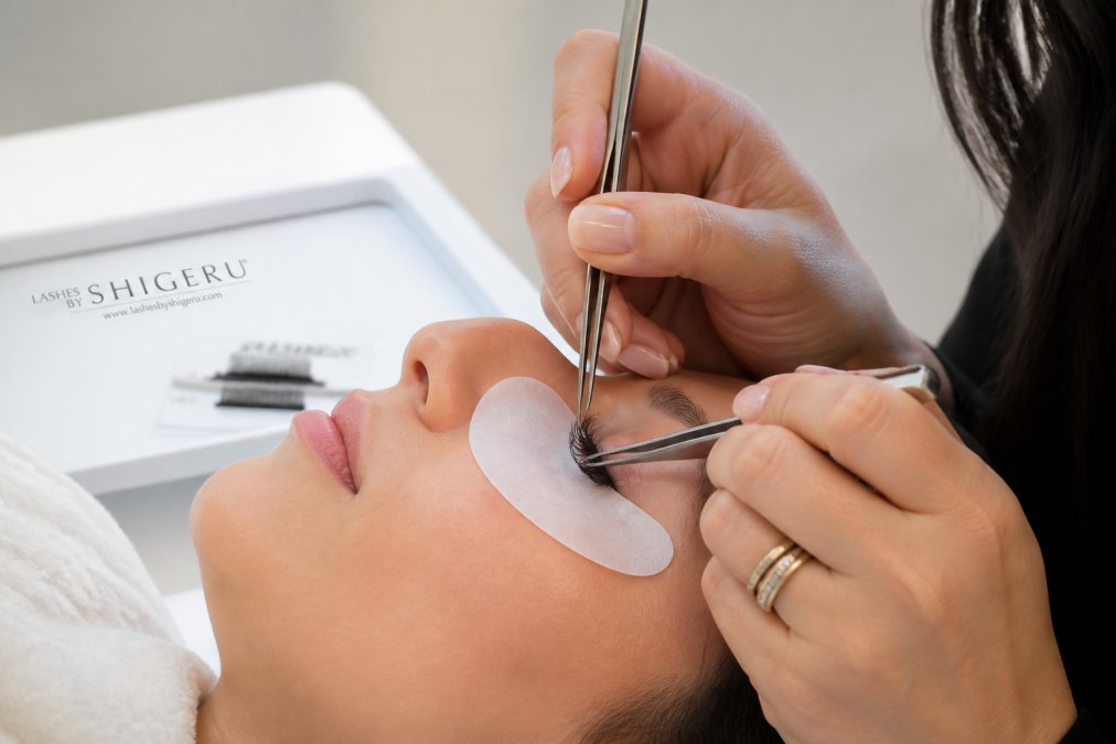 Close-up of a client's eye with professionally applied eyelash extensions showing natural curl and definition, with a trained lash technician's hands visible applying extensions under soft LED light in a clinical setting