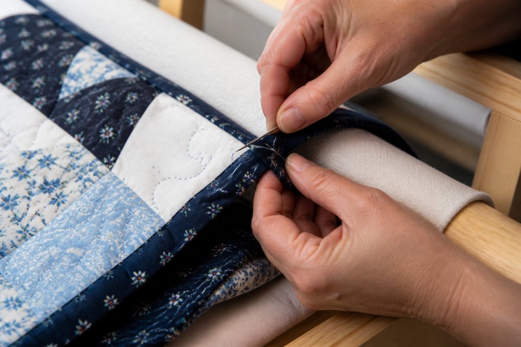 Close-up of hands carefully hand-stitching binding to the back of a finished quilt with coordinating fabric panels, showing the neat folded edge and detailed stitch work on a quilting frame