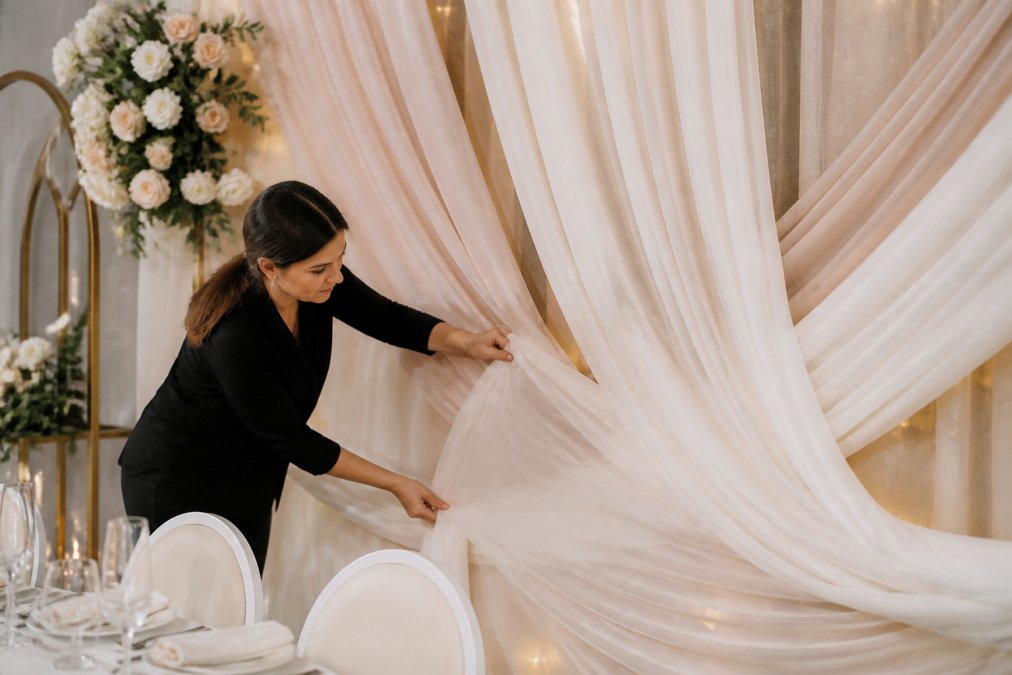 Flowing organza and chiffon fabrics draped elegantly against a wedding backdrop setup, hands adjusting the fabric to show shimmer and texture detail in soft natural light