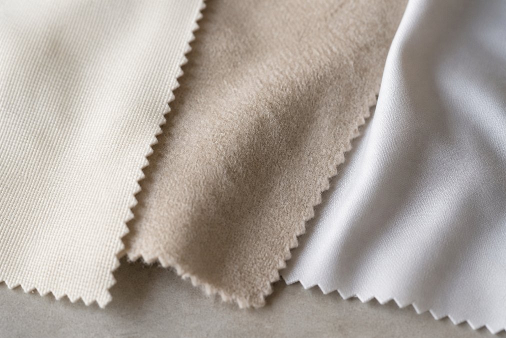Close-up of three different fabric swatches laid flat, cotton showing a tighter weave, soft brushed polyester with visible nap, and modal with a smooth lustrous finish, arranged on a neutral background with natural window lighting