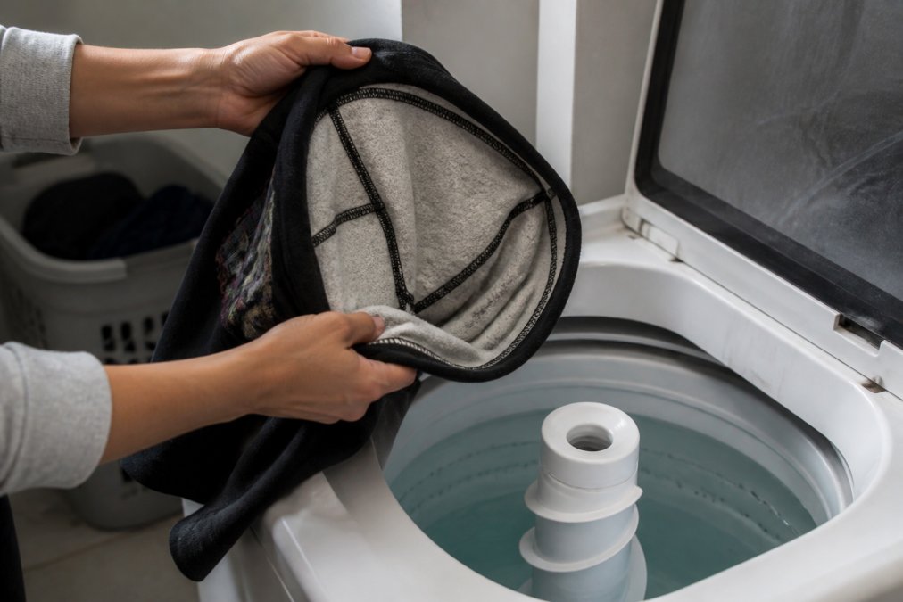 Hands gently turning a dark graphic hoodie inside out, revealing the interior seams and print backing, positioned above an open washing machine with cold water visible