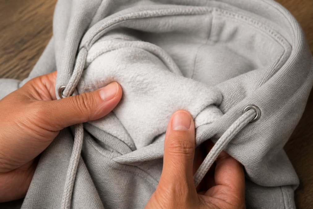 Close-up of a person's hands feeling the soft, brushed interior of a heather gray hoodie, showing the thick fleece texture and the double-lined hood with a color-matched drawcord