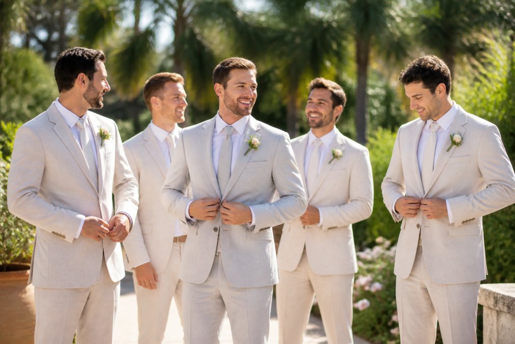 Groom and groomsmen wearing light-colored summer wedding suits while standing outdoors in a sunny garden venue
