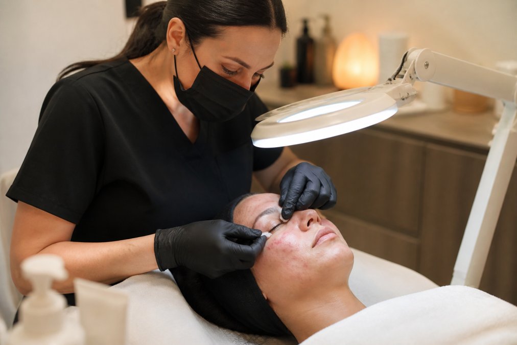 A licensed esthetician wearing gloves performing a facial extraction on a client in a clean, modern treatment room, using a magnifying lamp while the client rests on a treatment bed with soft, warm lighting