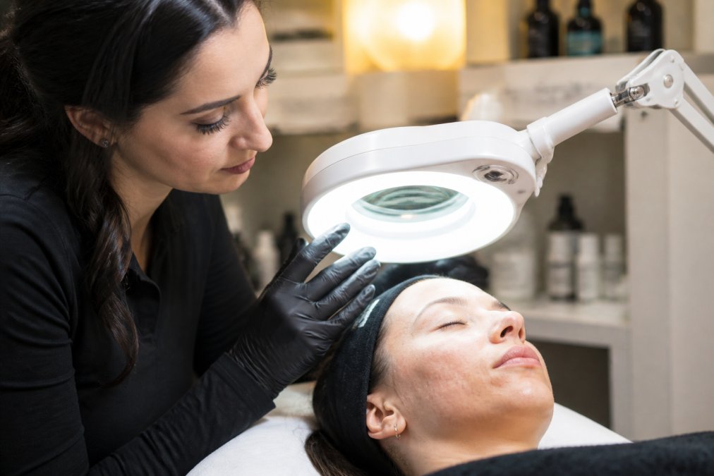 Close-up of a licensed esthetician examining a client's facial skin with a magnifying lamp in a clean, well-lit treatment room
