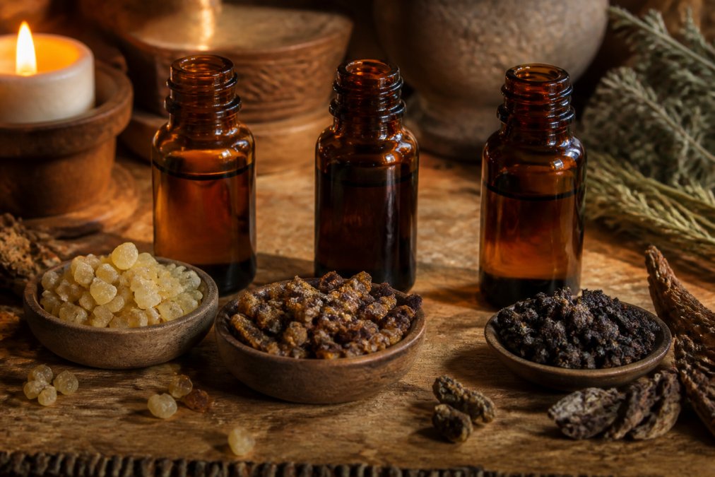 Amber glass bottles containing frankincense, myrrh, and spikenard essential oils arranged on a wooden surface with dried botanicals, candlelight creating warm shadows across the bottles