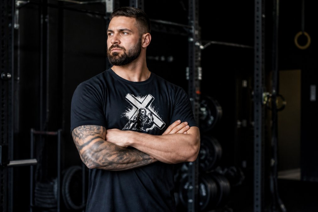 A fit man wearing a bold graphic faith-based cotton t-shirt in a gym setting, arms crossed, confident posture, dramatic lighting against a dark background