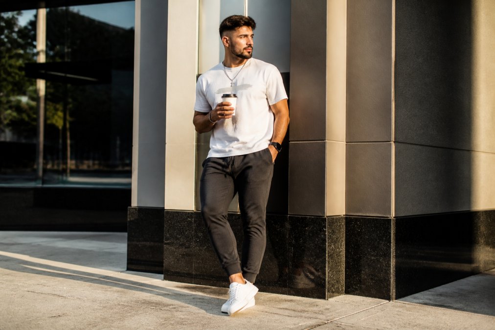 A man in a clean faith-inspired graphic tee, joggers, and white sneakers holding a coffee cup outside a modern building in morning light, relaxed streetwear posture that reads casual and confident