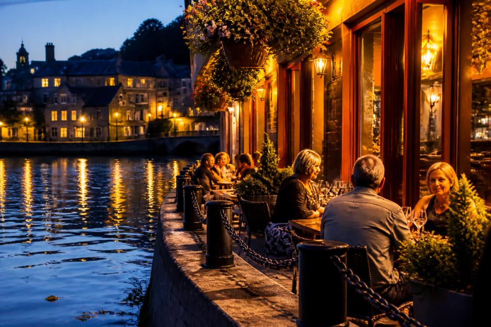 Evening scene at a riverside bistro with warm lighting reflecting off water, diners seated at tables with historic stone buildings visible in the background, soft amber glow from interior creating intimate atmosphere