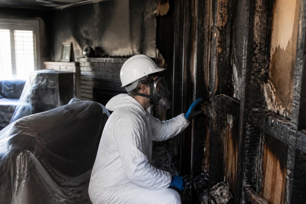 A restoration technician in protective gear and respirator inspecting charred wall framing inside a fire-damaged home, with soot-stained walls and salvageable furniture covered in plastic sheeting nearby