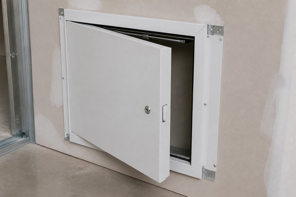 Close-up of a properly installed fire-rated access door with visible self-closing mechanism and gasket seal in a commercial wall assembly, showing frame anchoring and drywall integration